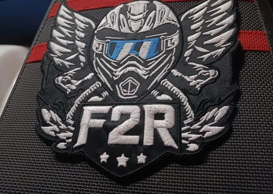 Grand écusson brodé : un casque de motocross ailé et le sigle F2R, en fil blanc et gris sur fond noir texturé.